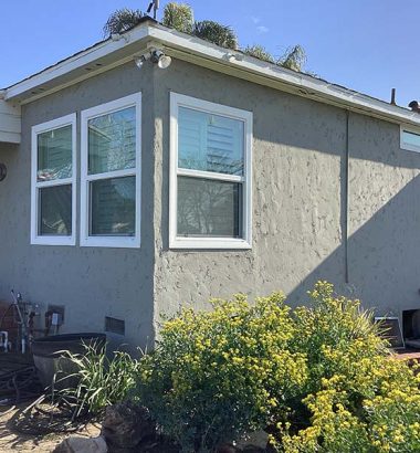A gray house exterior with multiple windows, bordered by lush green foliage and yellow flowers under a clear blue sky.