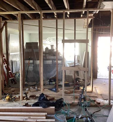 Construction scene showing two workers framing a wall, with tools and materials scattered in a partially renovated room.