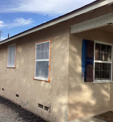A person on a ladder paints a house's exterior, with windows covered and part of the wall in blue, under a blue sky.