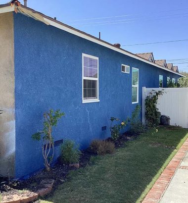 A bright blue house exterior with a garden, contrasting with a beige side. It features windows and a gated area in the background.