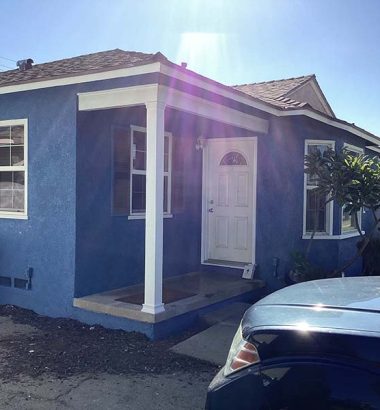 A blue house with white trim features a front porch, a white door, and nearby greenery, with a trash can visible on the left.