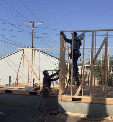 Two men constructing the wooden frame of a house, focused on their work with tools and materials around them.