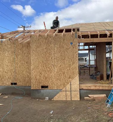 A house under construction with a man working on the roof, surrounded by scaffolding and building materials.