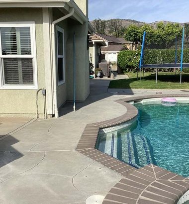 A swimming pool surrounded by a tiled patio, with a house visible in the background.