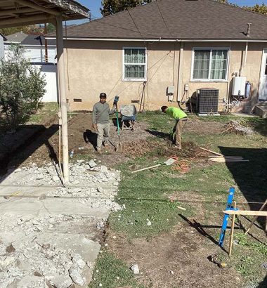 Two men are landscaping a backyard in a residential neighborhood, surrounded by grass and trees.