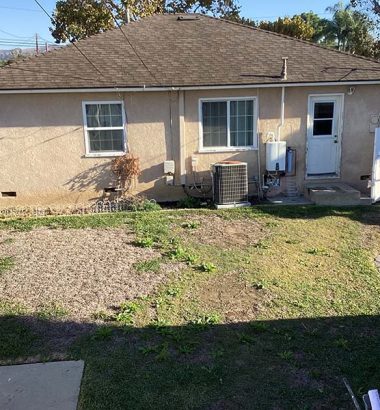 A small house surrounded by a modest yard, featuring grass and a few plants, under a clear blue sky.