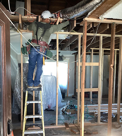Worker on a ladder adjusts ductwork in a partially constructed room, surrounded by exposed framing and construction materials.