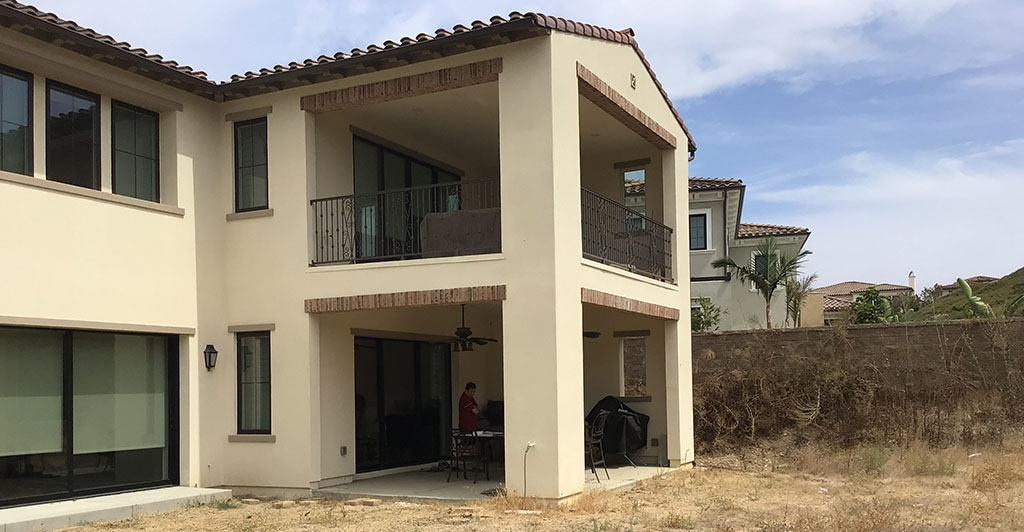 A house featuring a balcony above and a patio area below, surrounded by greenery.
